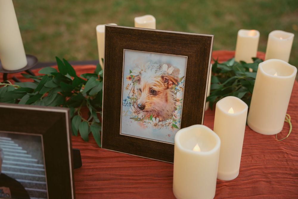 A framed photograph sits on a red tablecloth surrounded by white candles and greenery.