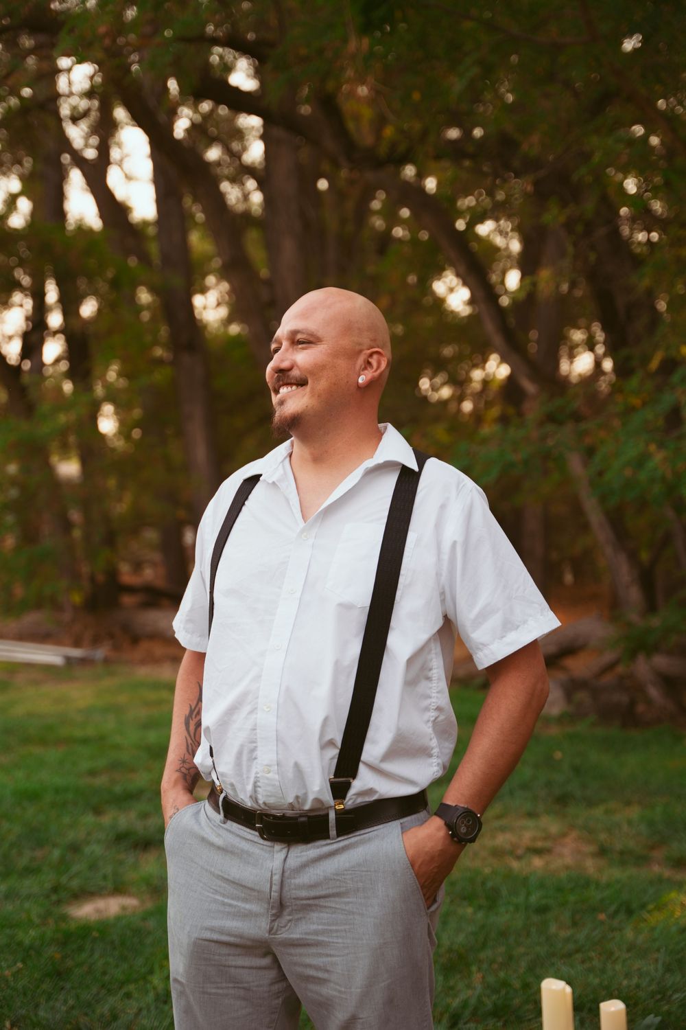 A person in suspenders and white shirt smiles while standing in a wooded outdoor setting.