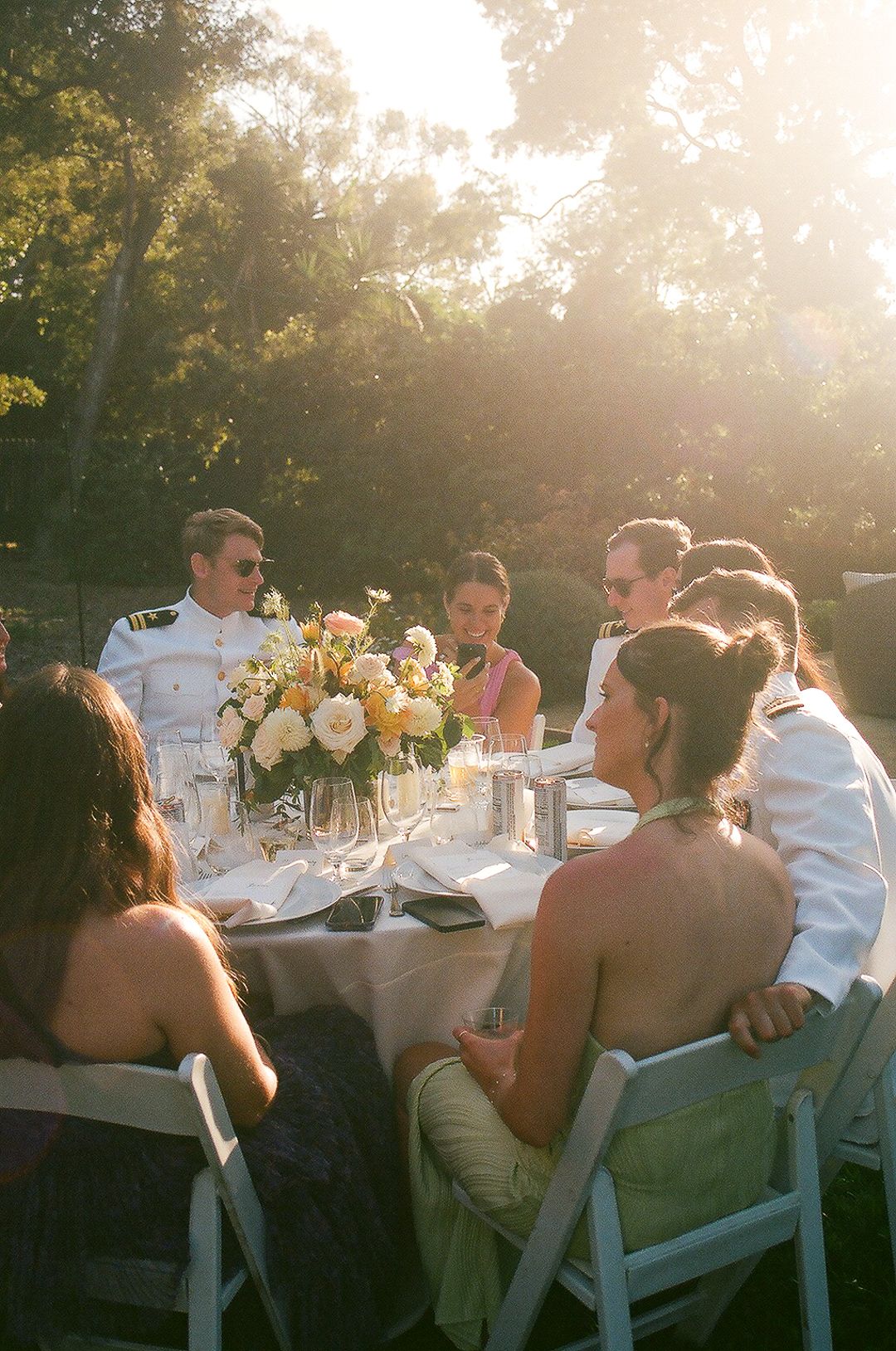 Outdoor dinner gathering at sunset with elegant table settings and yellow floral centerpieces bathed in golden light.