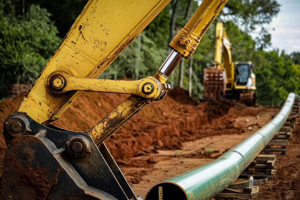 Yellow excavator arm installing large industrial pipeline in dirt trench.