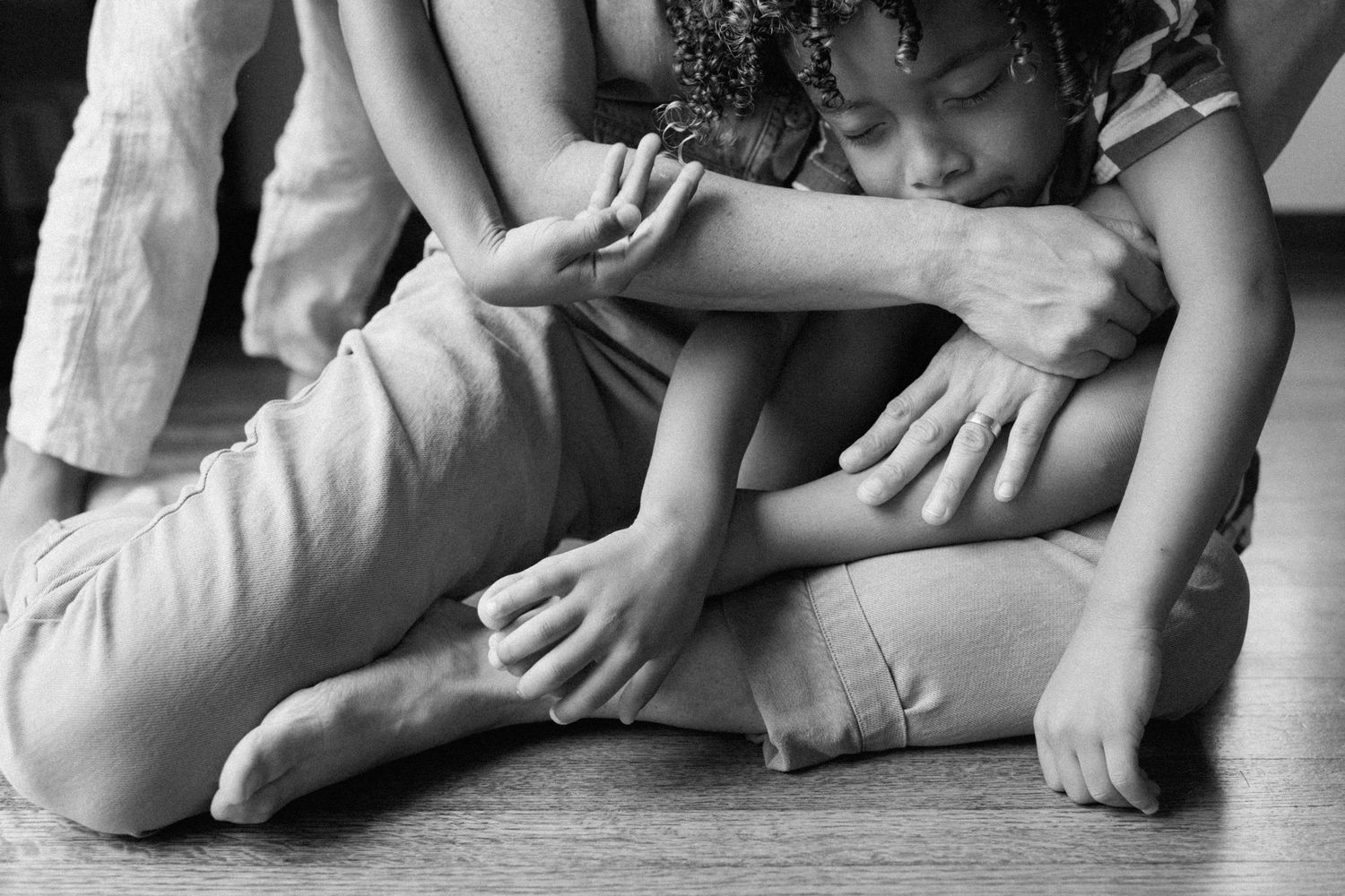 A black and white close-up photograph shows hands and feet resting together on a wooden floor.