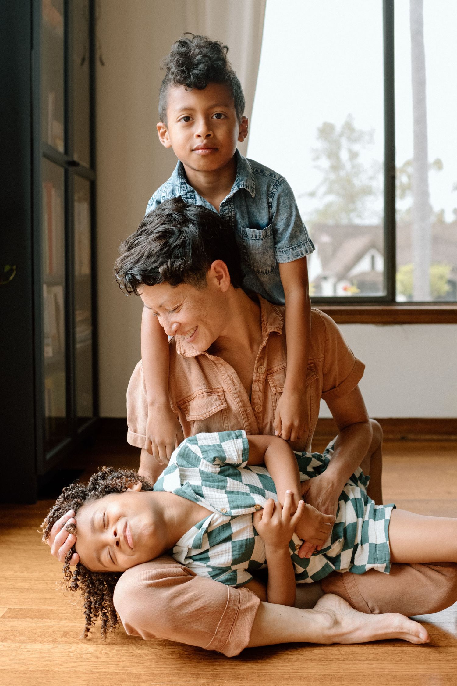 Family playfully wrestling and laughing together on a hardwood floor near a window in natural lighting.