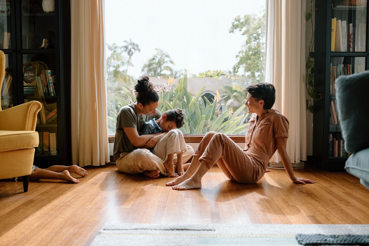 Two people sit on a hardwood floor by large window having conversation in cozy living room with natural light.
