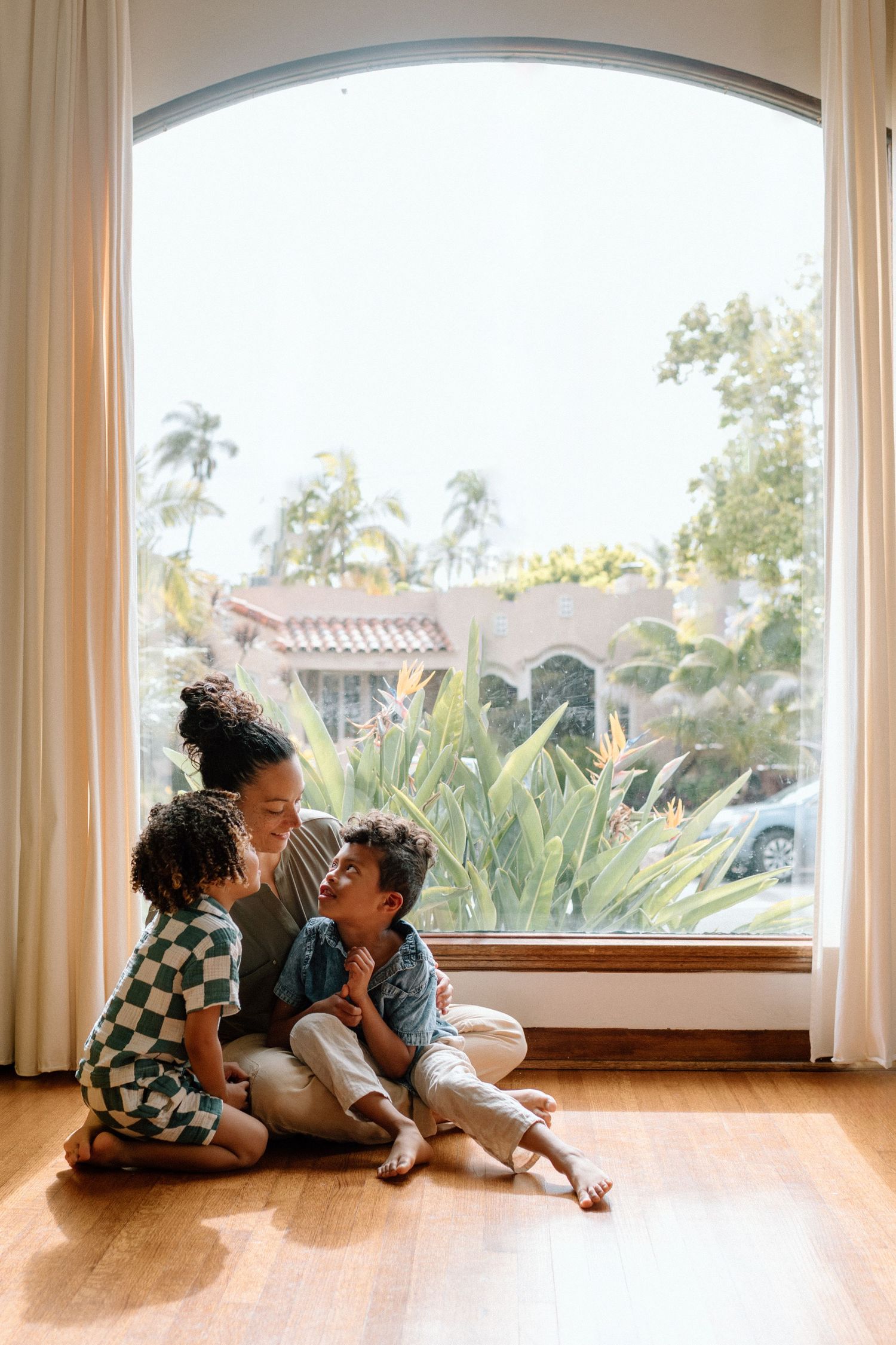 Three connected images show a family lying together on a hardwood floor by large windows in a sunlit room.