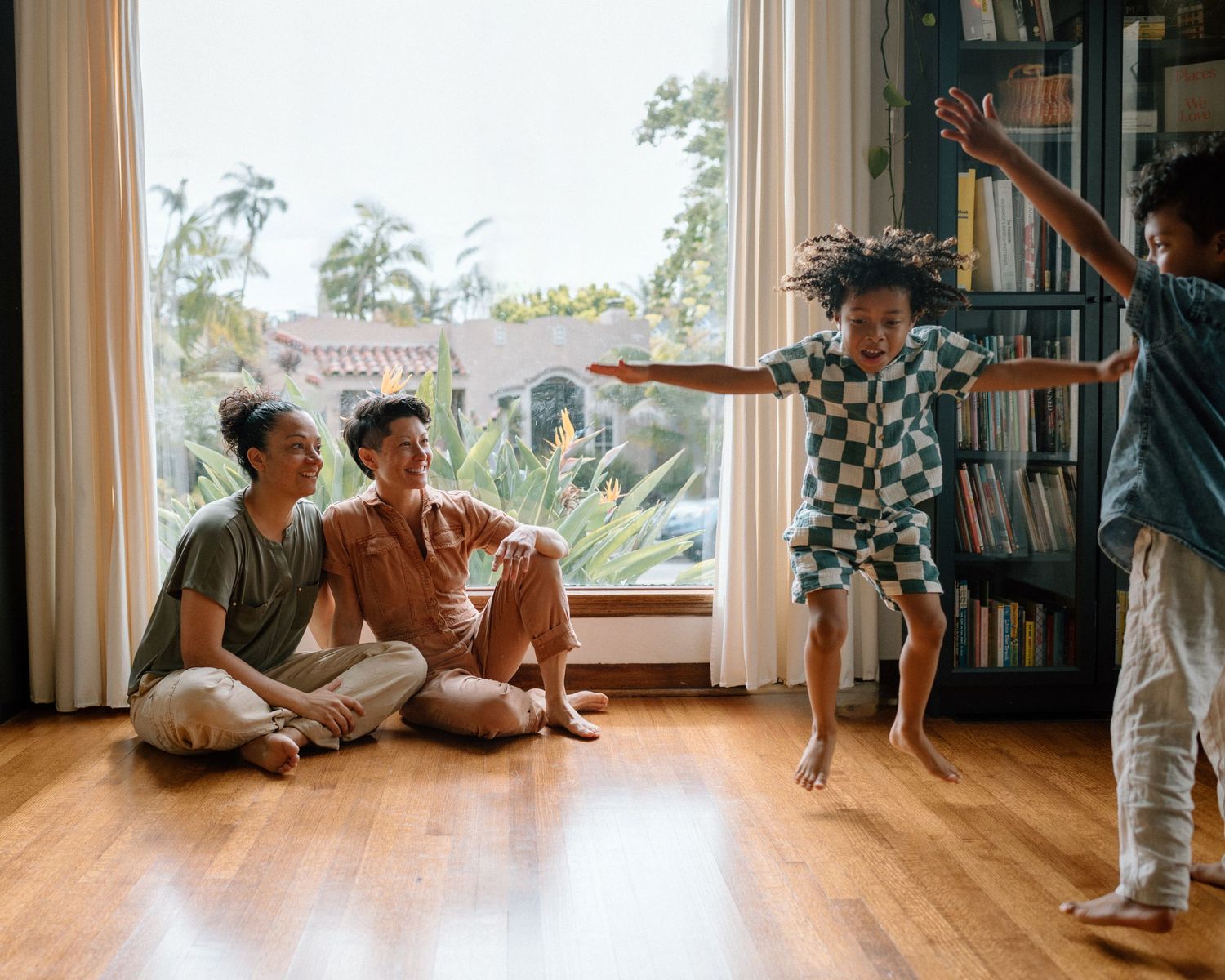 Four people enjoy a playful moment together in a sunny living room with wood floors and window views.