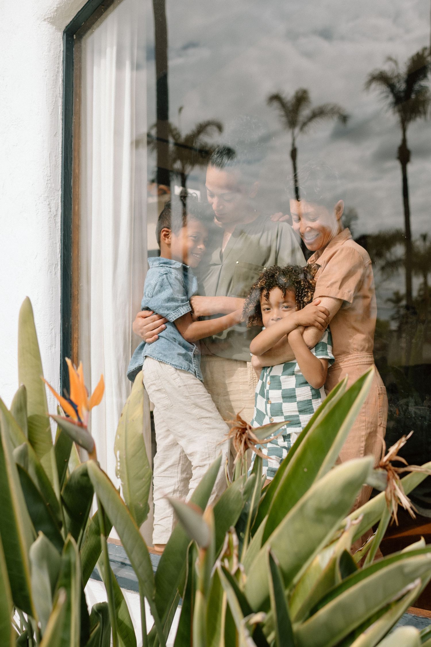 A family gazes through a window together, framed by tropical palm trees and bird of paradise plants in the foreground.