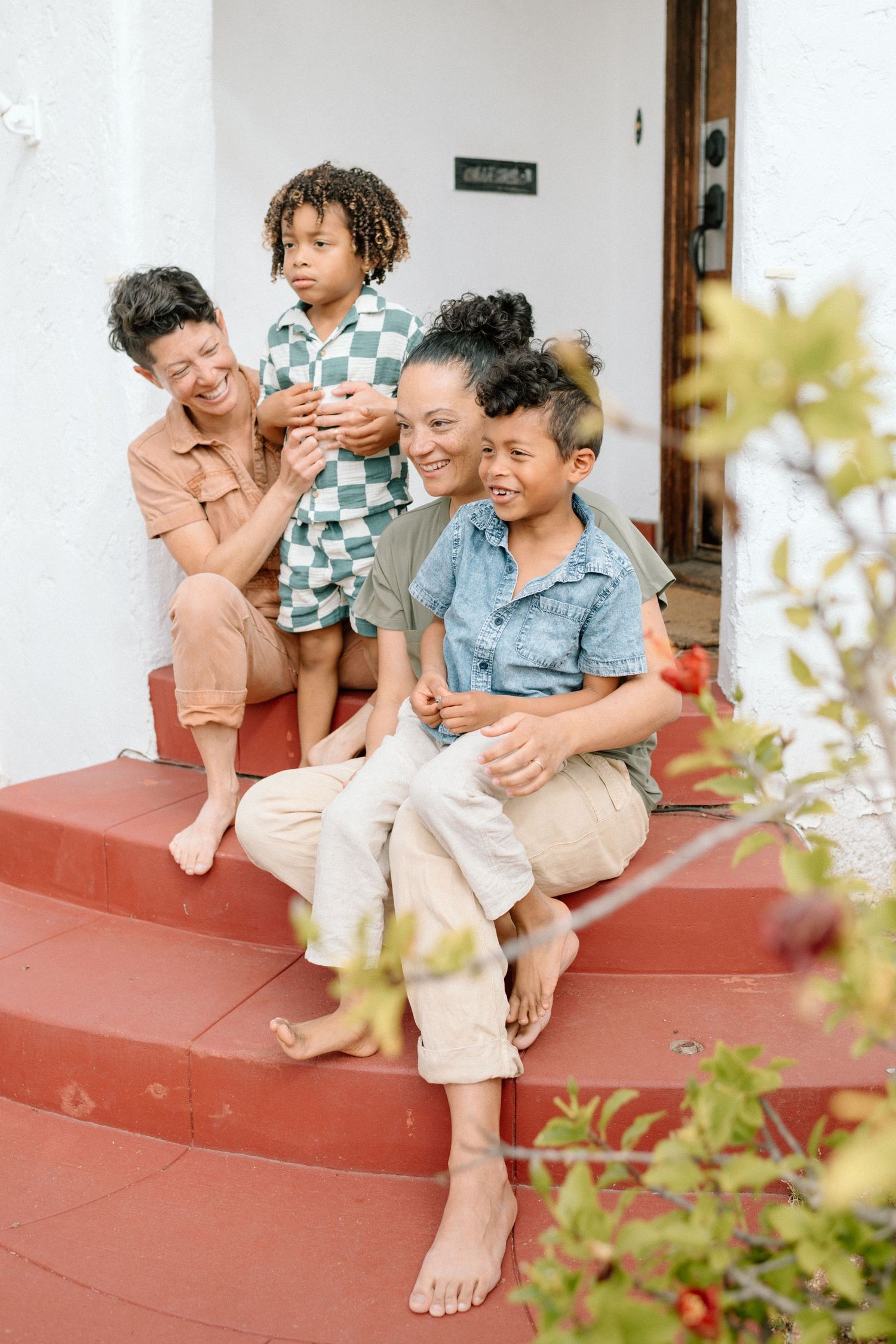 Family sitting together barefoot on red front porch steps sharing a joyful moment on a sunny day.