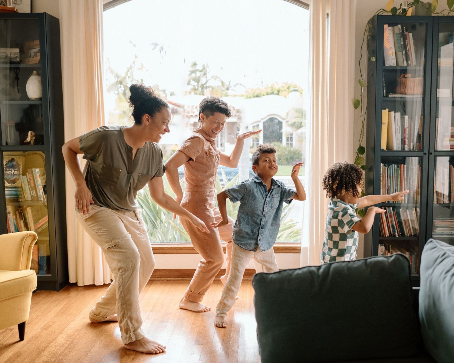 A joyful group dances together in a sunlit living room with bookshelves and a dark couch.