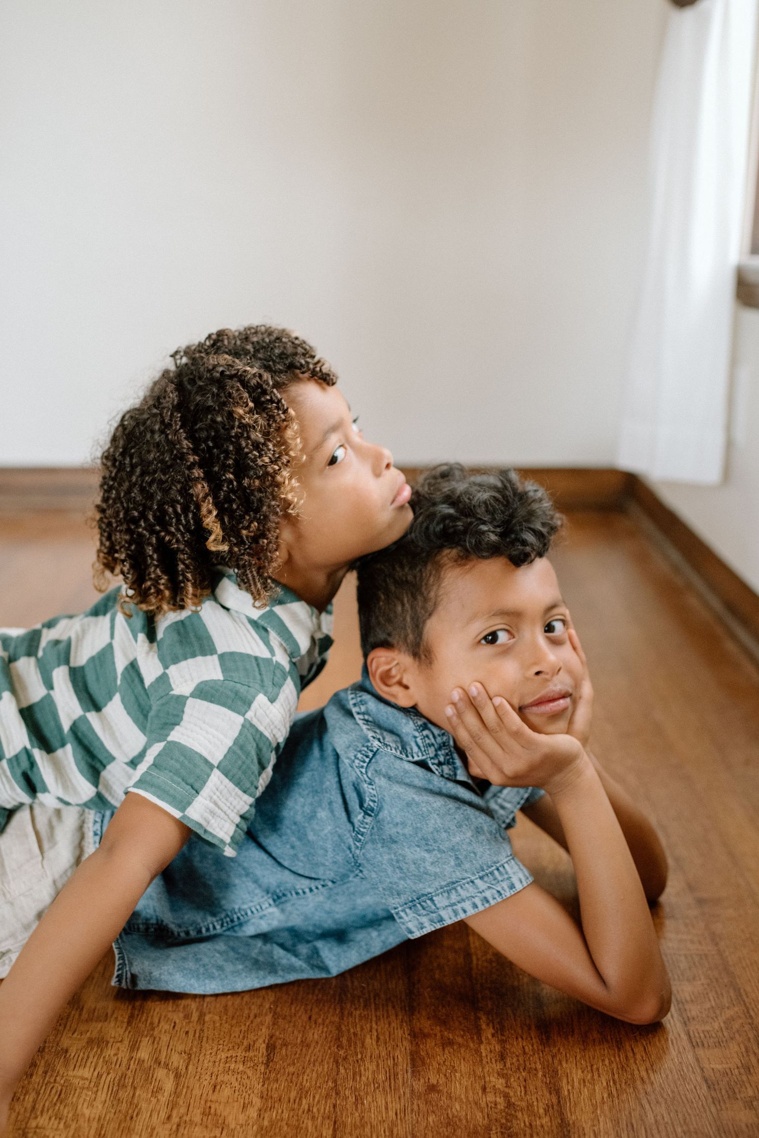Two children lie on a hardwood floor making playful expressions, one wearing a checkered shirt and one in denim.