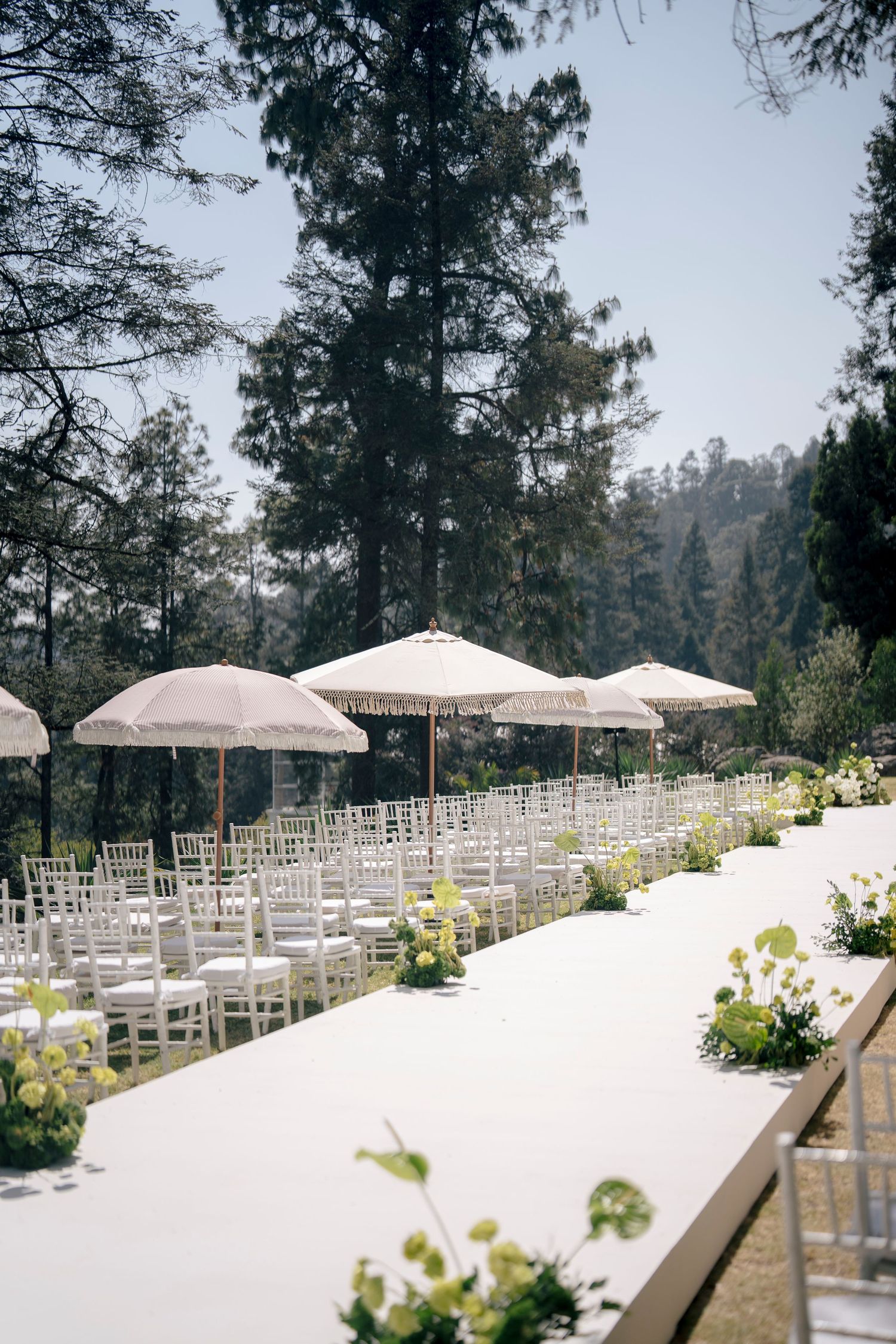 White chairs and umbrellas line an outdoor wedding aisle set in a forest with mountain views and floral arrangements.