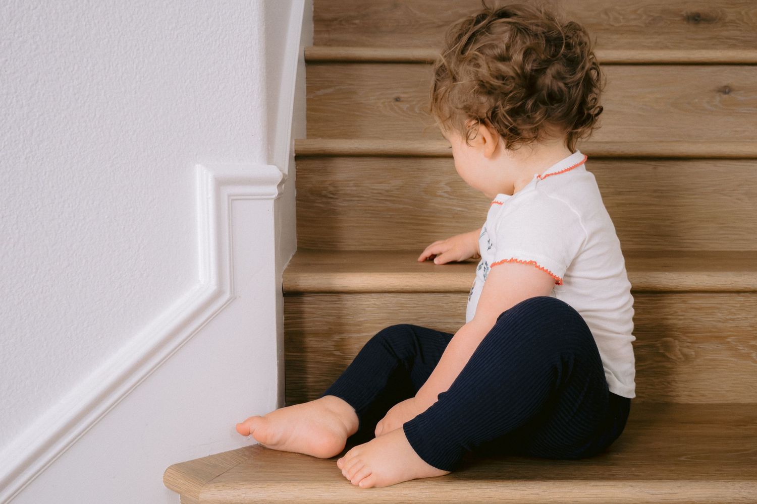 A toddler in a white shirt and navy blue pants crawls up wooden stairs.