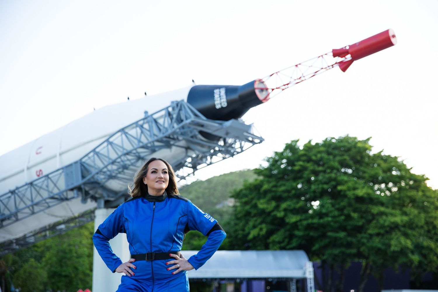 Someone in a bright blue outfit stands below a large bottle-shaped sculpture or art installation outdoors.