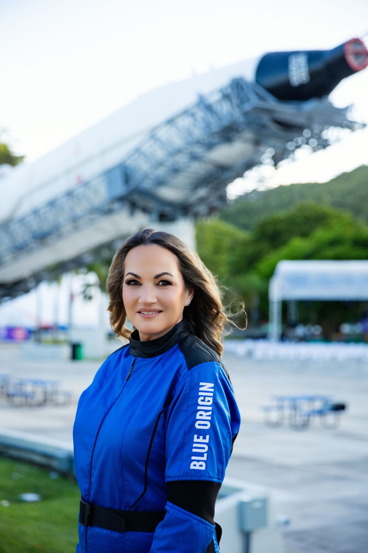 A person in a bright blue flight suit stands outdoors near some seating and structures during an event.