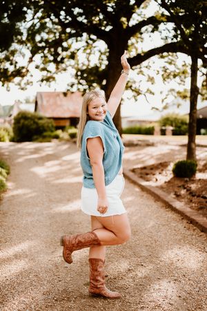 A playful figure in a blue top and brown boots dances joyfully along a dirt path under trees.