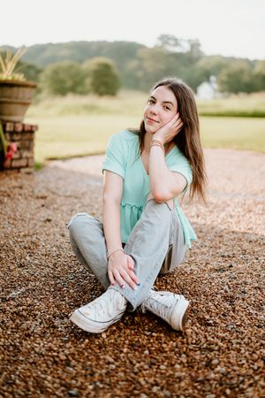 A person in a mint green top and gray pants sits casually on a gravel ground while looking away thoughtfully.
