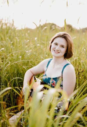 Someone in a teal dress sits among tall grass in a sunlit field during a summer photoshoot.