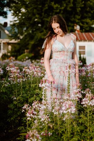 Figure in a flowing pastel dress stands among pink garden flowers during golden hour at a rustic estate.