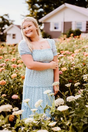 A person in a light blue dress poses among blooming white and pink zinnia flowers in a summer garden setting.