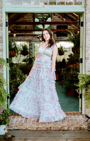 A flowing floral dress in soft blue and pink creates an ethereal look against a greenhouse doorway filled with lush plants.