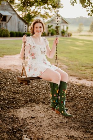 A person in a floral dress and green boots swings joyfully at sunset in a rustic outdoor setting.