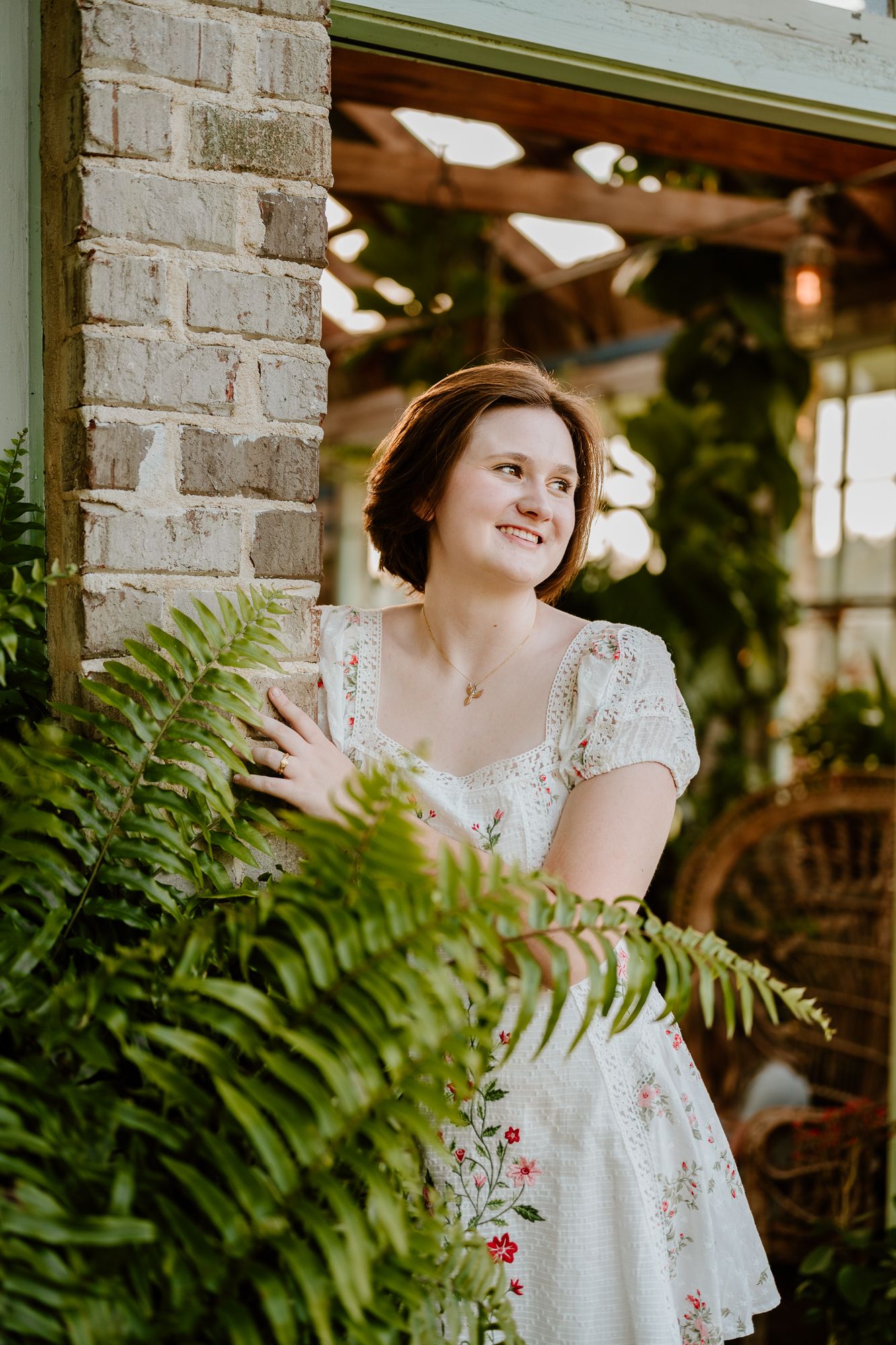 Woman in a white lace dress poses near green ferns and exposed brick wall in an indoor garden setting.