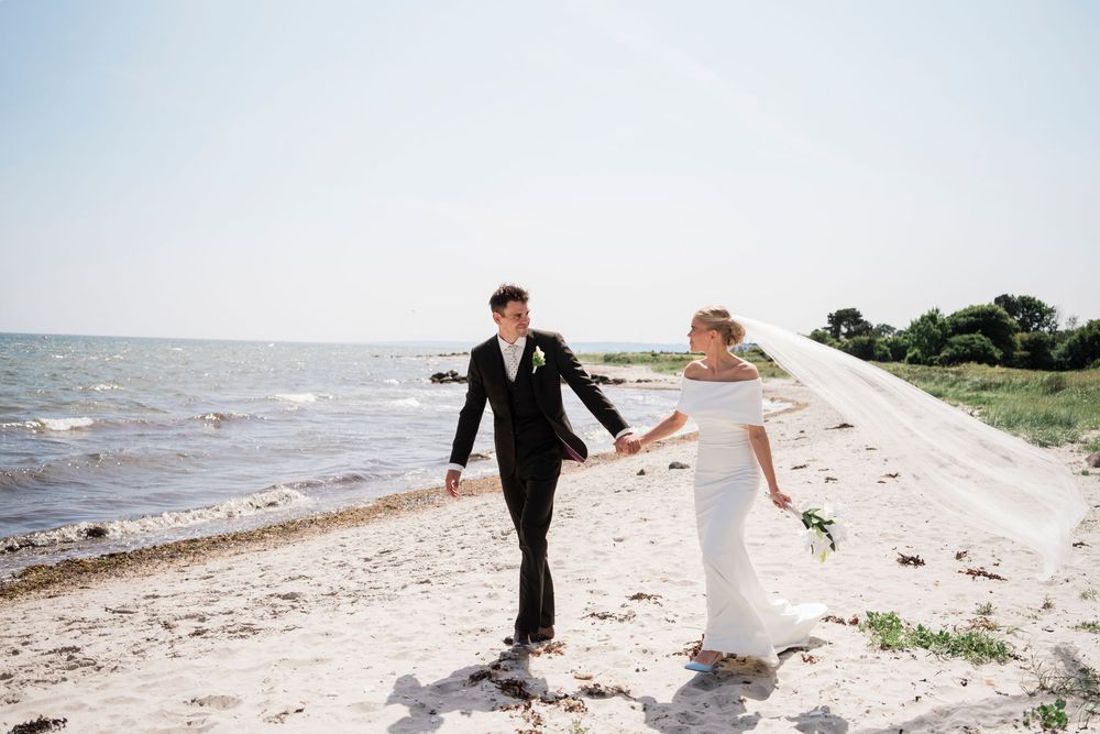 A couple in wedding attire strolls hand in hand along a scenic beach on a bright sunny day.