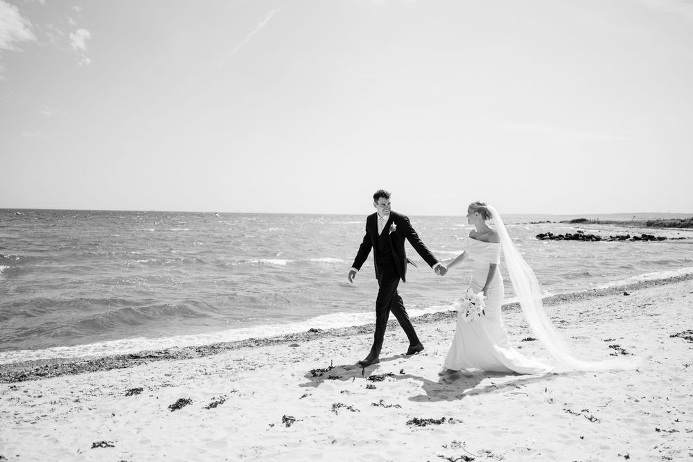 A romantic couple in formal attire runs playfully along a sandy beach at sunset.
