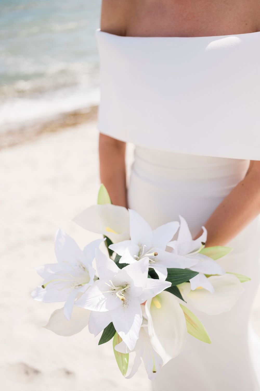 A white lily bridal bouquet held against an elegant off-shoulder wedding gown on a sandy beach.