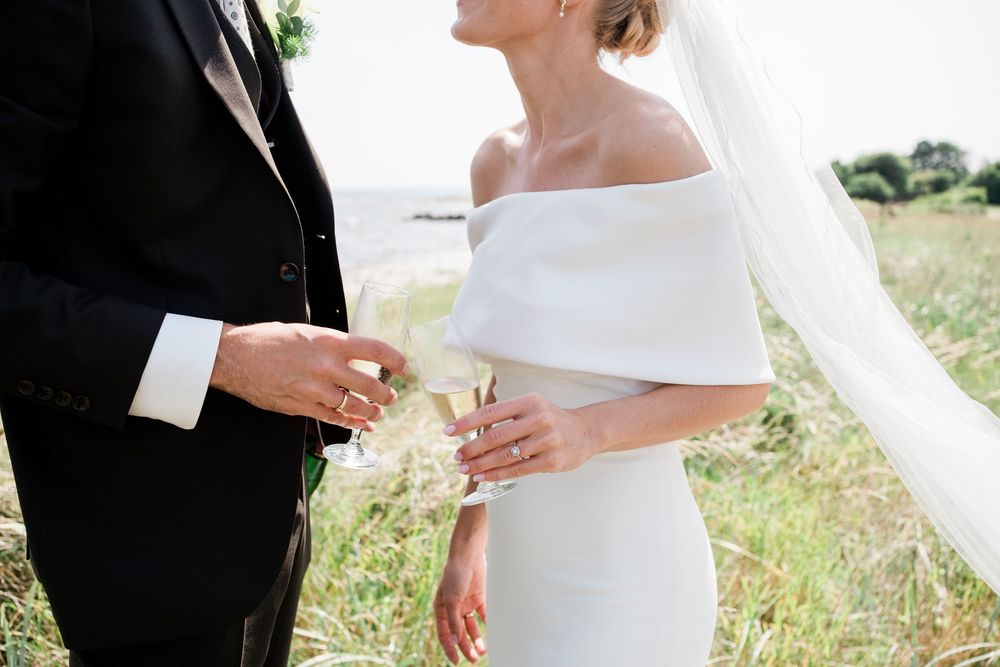 A romantic wedding couple holds hands in nature, with focus on their intertwined fingers against sunlit grass.