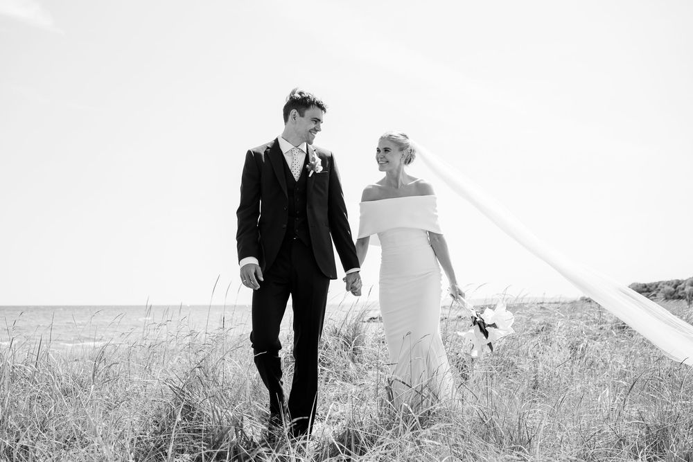 A couple in formal wedding attire walking together through a grass field captured in timeless black and white photography.