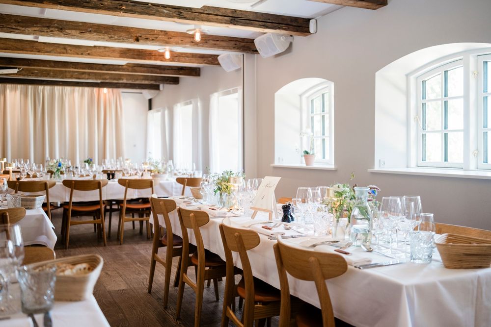 Contemporary restaurant dining room with exposed wooden beams and elegant place settings on white tablecloths.