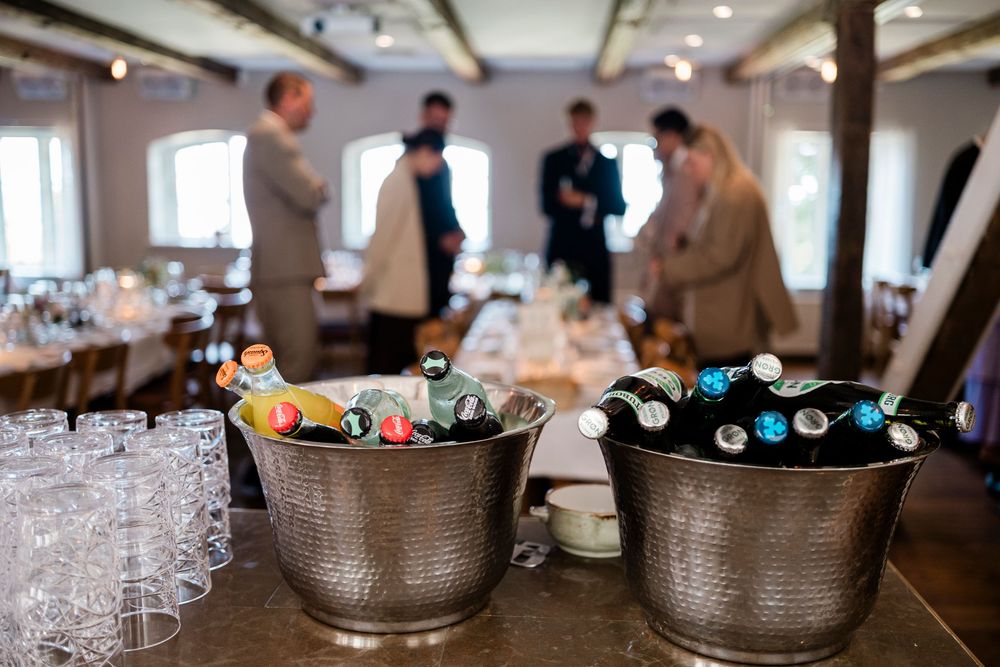 Two silver ice buckets filled with assorted beverages sit on a table at an indoor event with guests mingling in background.