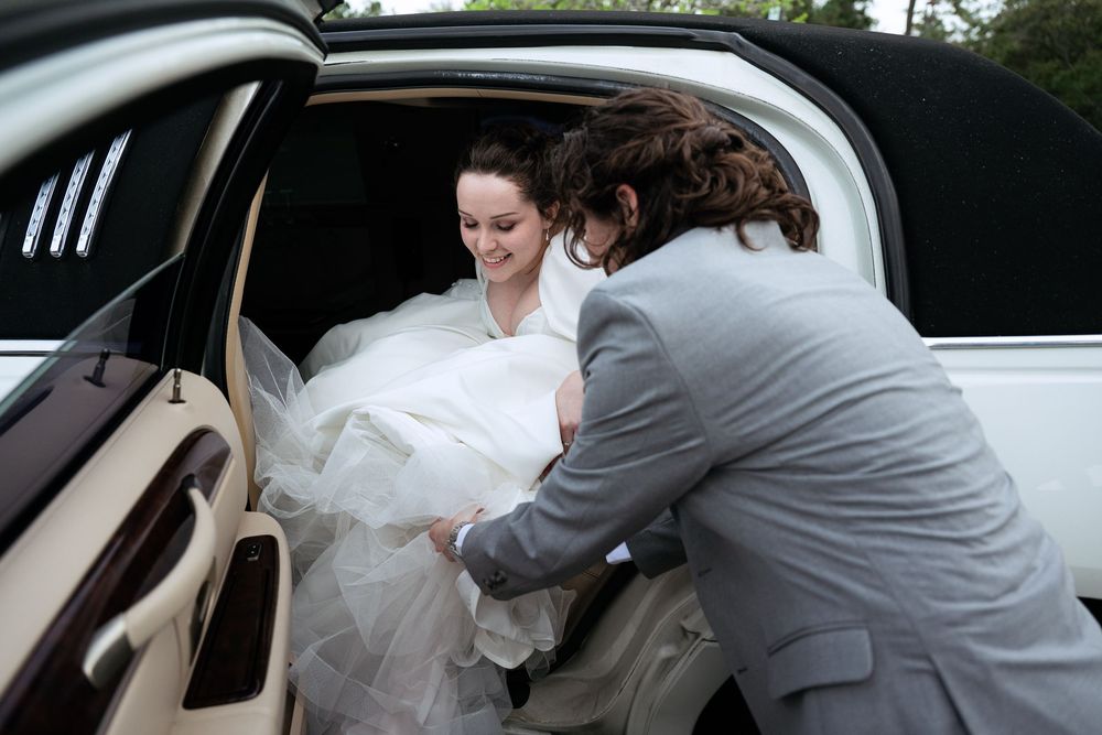 A bride in a white wedding dress is being assisted into a car by someone wearing a gray suit.