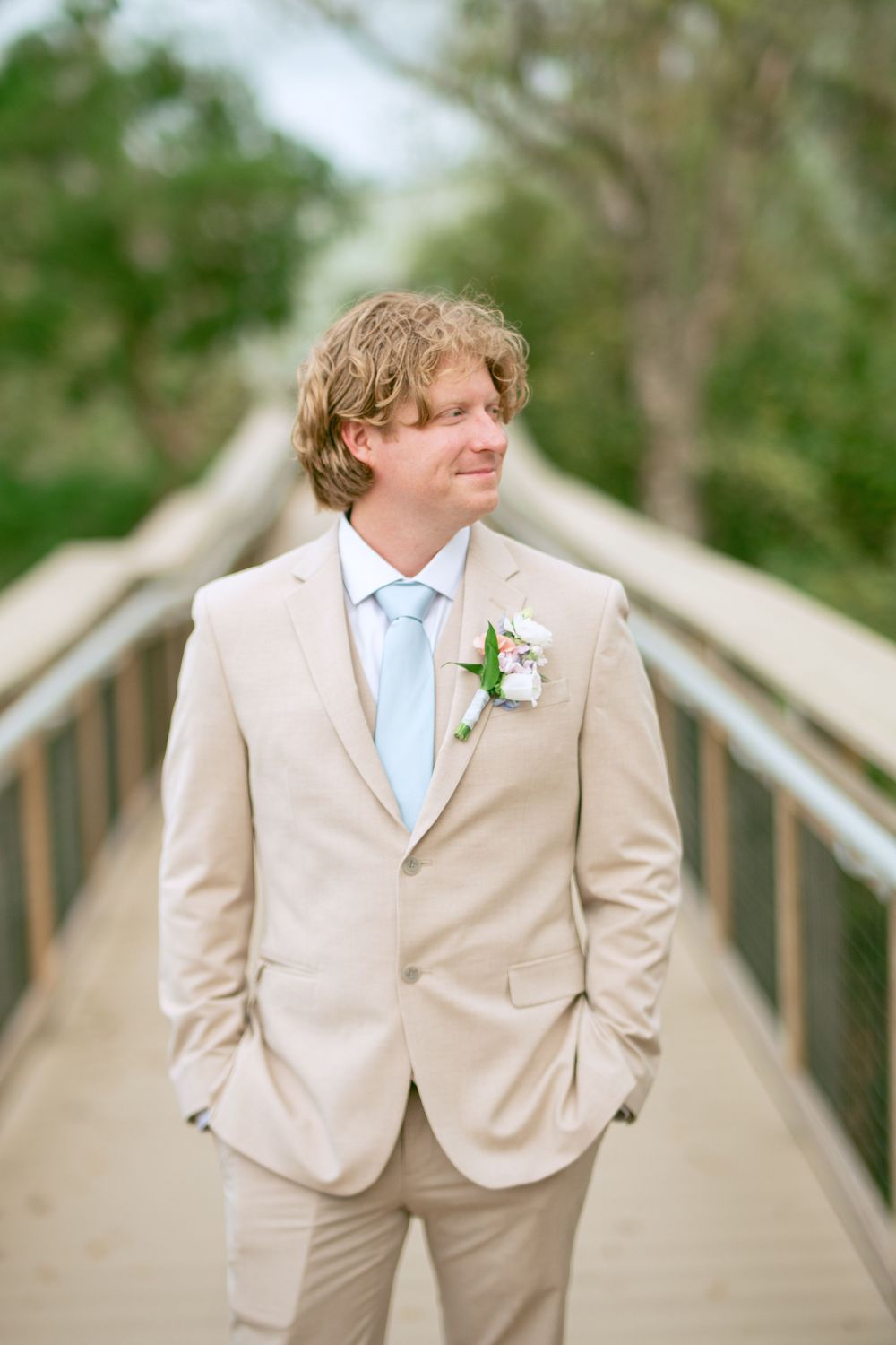 Groom in light beige suit with blue tie poses on wooden bridge walkway.