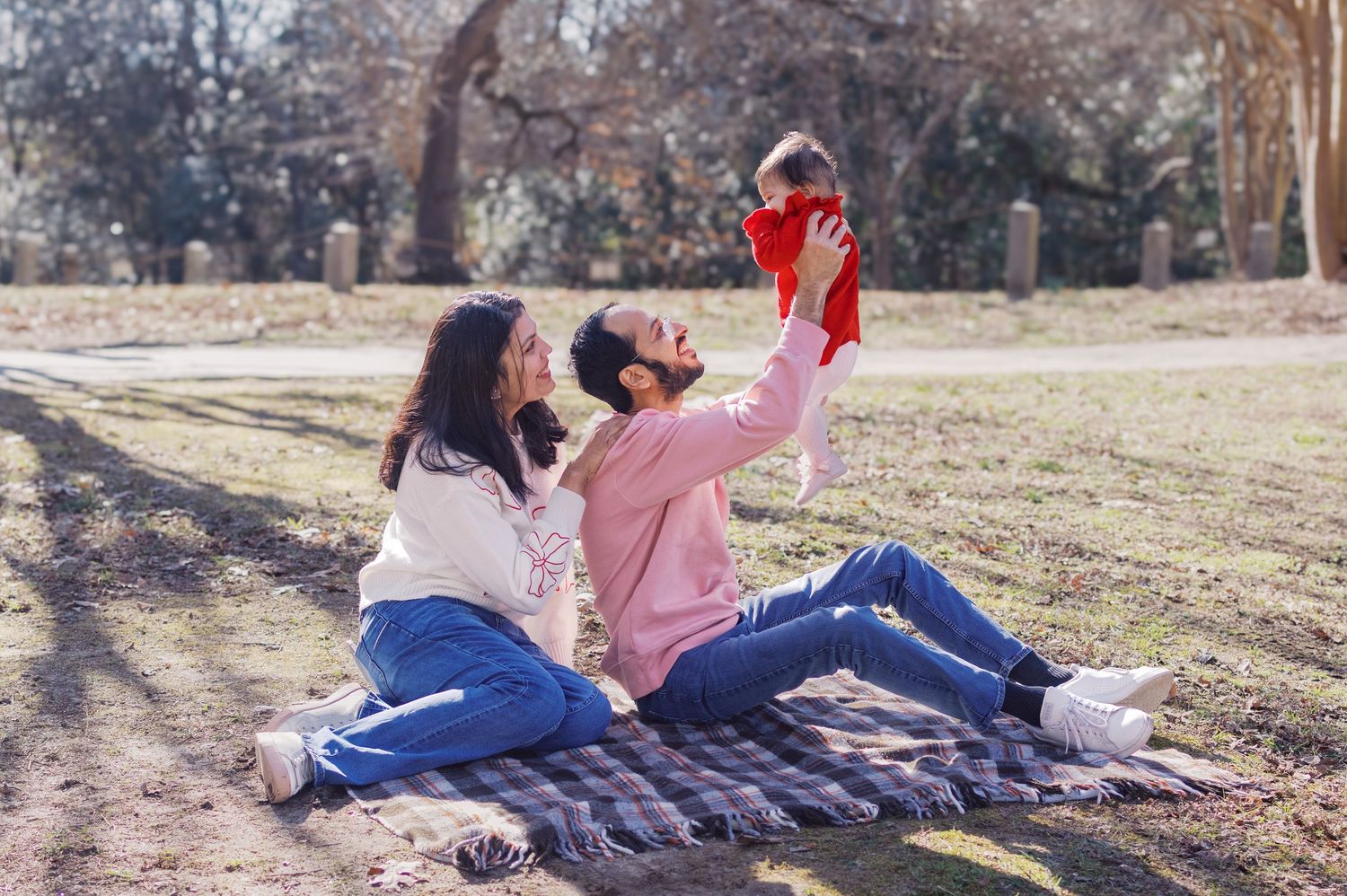 A couple lying on a picnic blanket plays with their baby in a sunny park.
