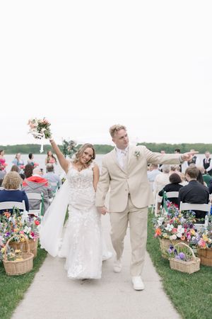 Newlyweds joyfully walk down aisle at outdoor wedding ceremony with floral decor and seated guests.