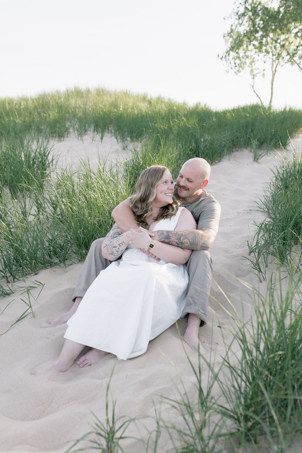 A couple shares an intimate moment while sitting together on sandy dunes surrounded by tall beach grass.