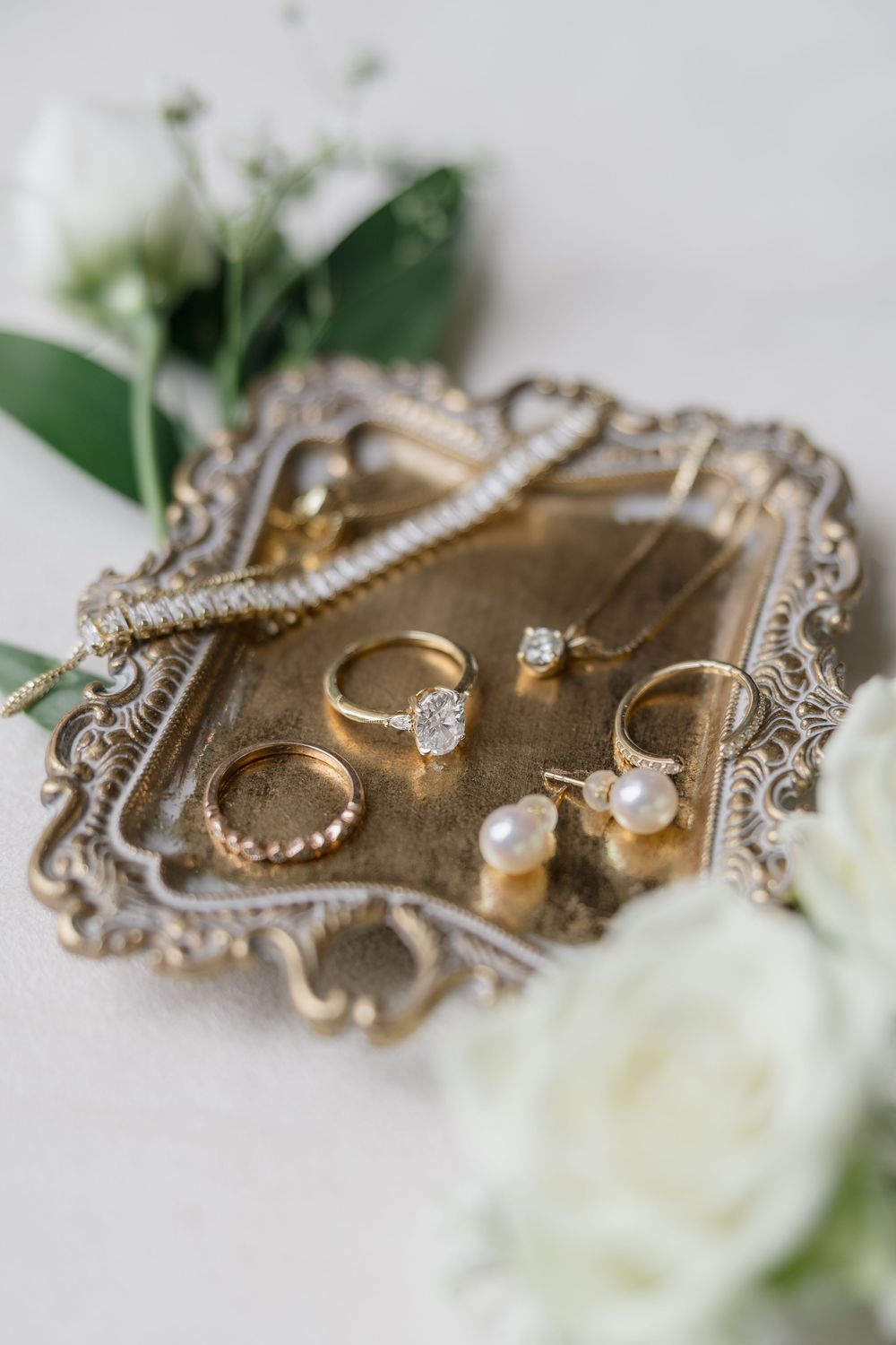 Gold jewelry rings with diamonds and pearls displayed on an ornate vintage silver tray with white flowers.