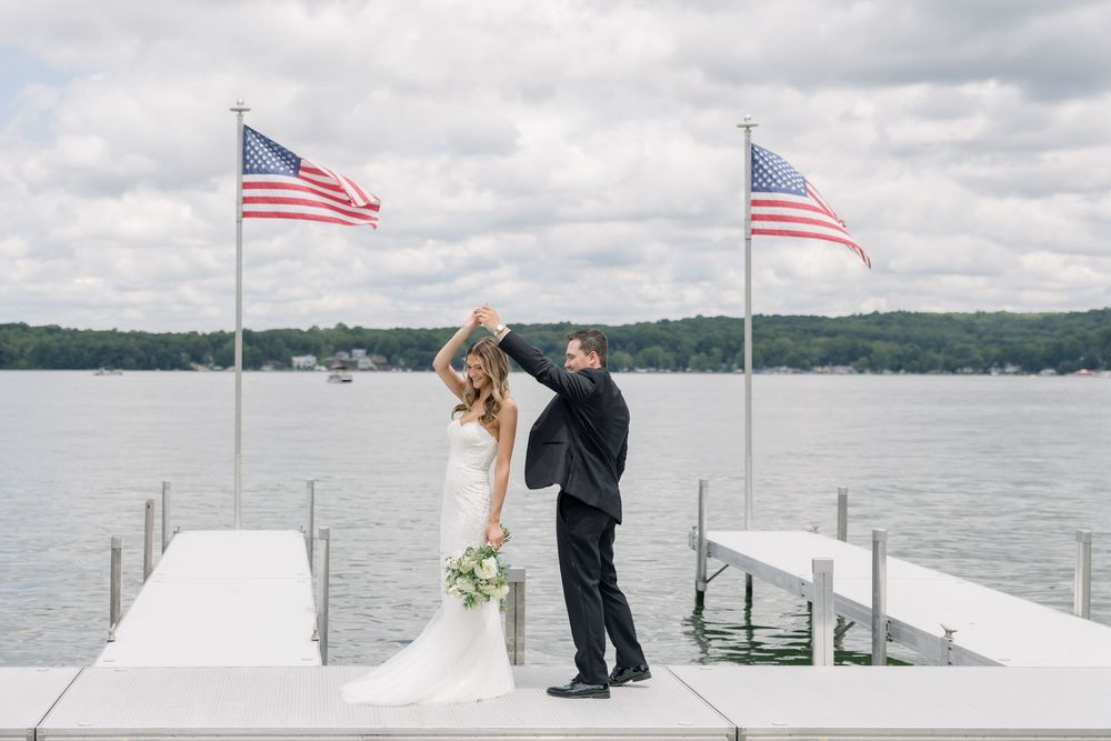 Two figures in wedding attire stand on a dock with American flags and a lake view in summer.