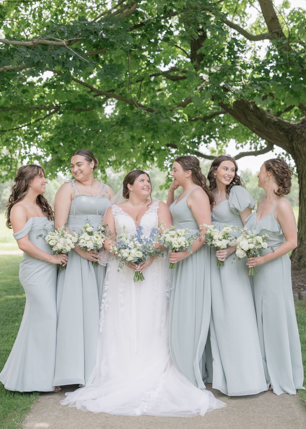A bridal party in flowing light blue bridesmaid dresses and white wedding gown pose together under a leafy green tree.