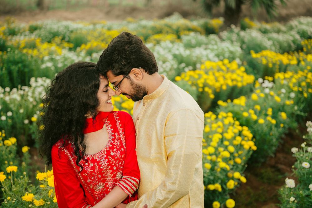 A romantic couple in red and cream colored outfits embrace in a field of blooming yellow flowers.
