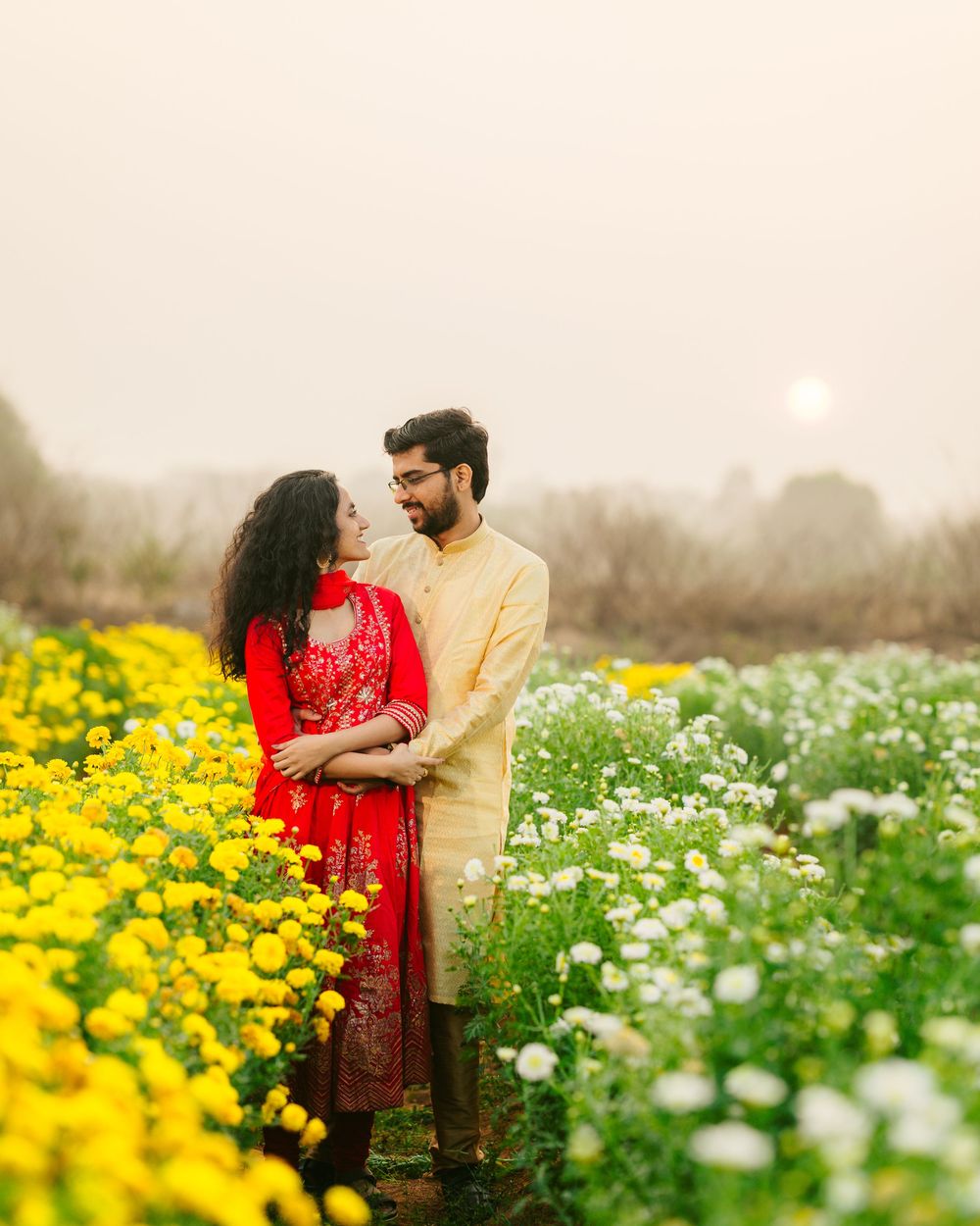 A romantic couple stands amid vibrant yellow and white wildflowers in a misty field.