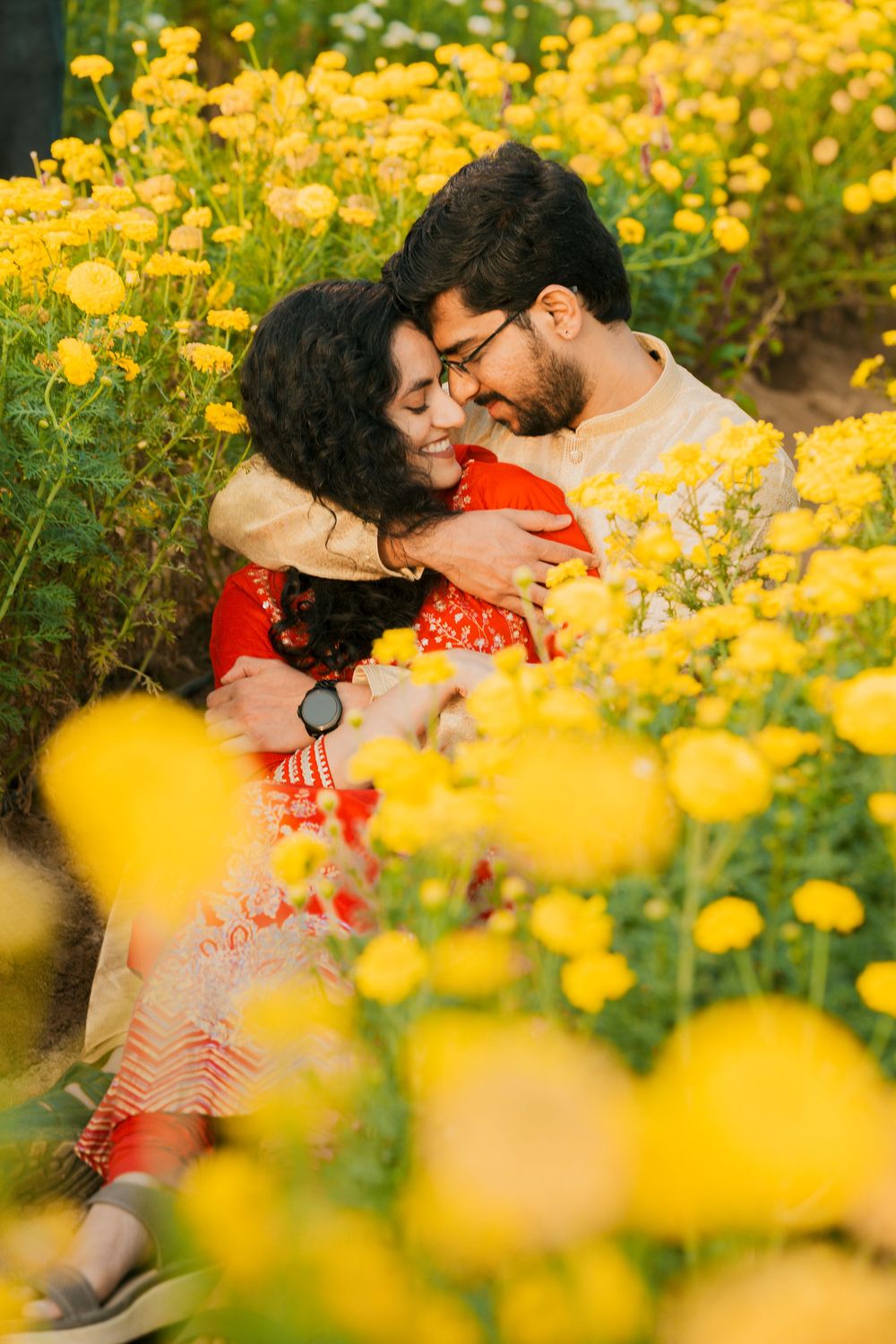 A couple embraces amid a garden of vibrant yellow flowers while wearing traditional Indian clothing.