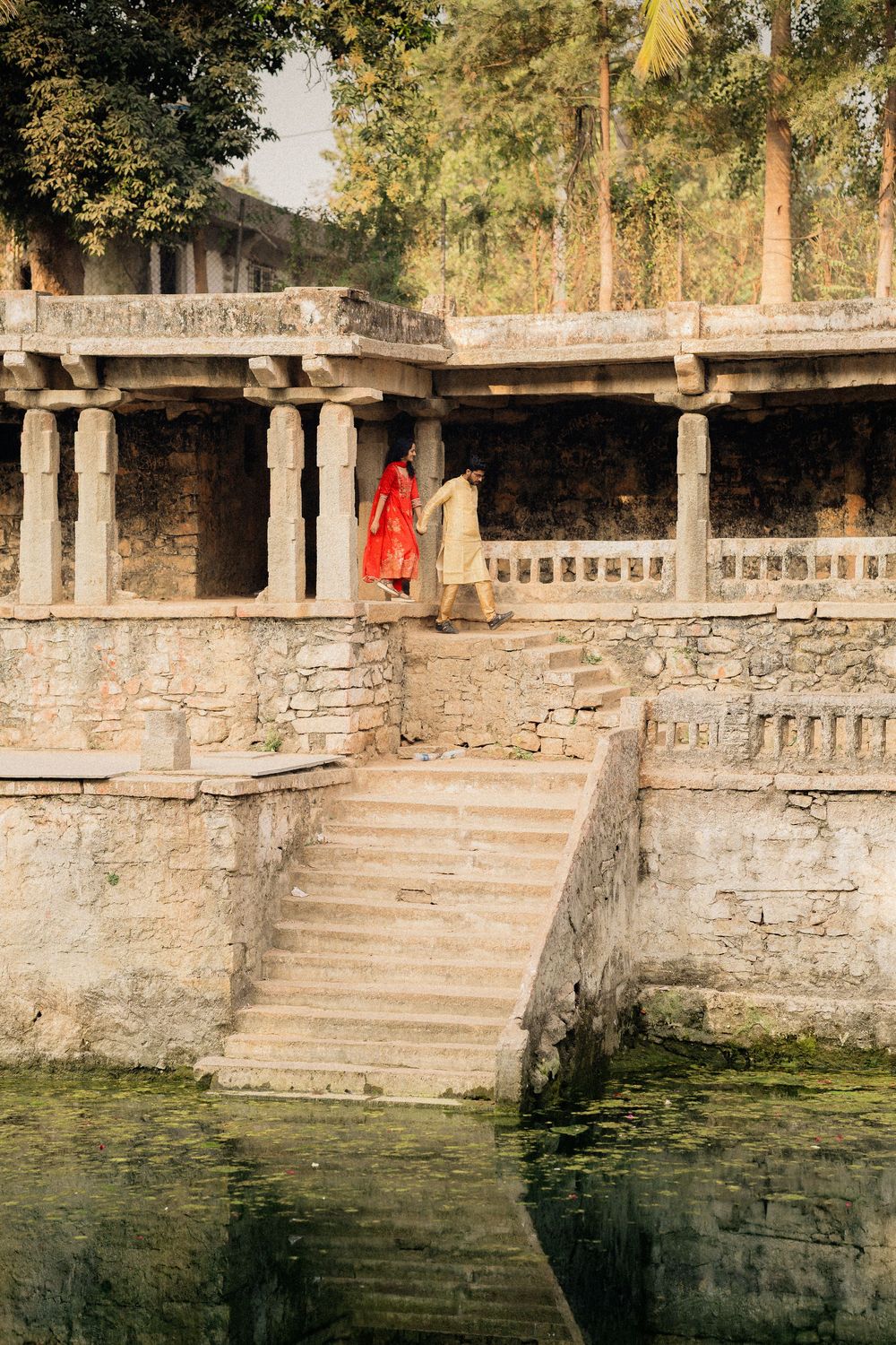 Ancient stone steps lead down to a tranquil pond with stone walls and a bright red garment hanging in the sunlit background.