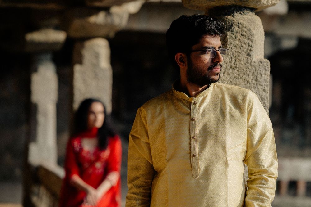A man in yellow kurta stands in focus at a historic temple column with a woman in red in background.