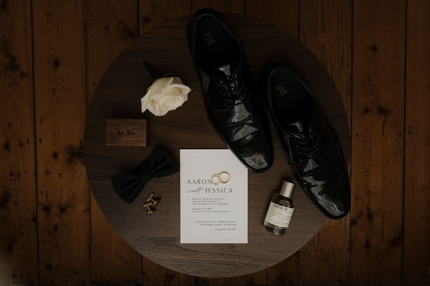 Wedding details including dress shoes, bow tie and invitation on wooden surface.