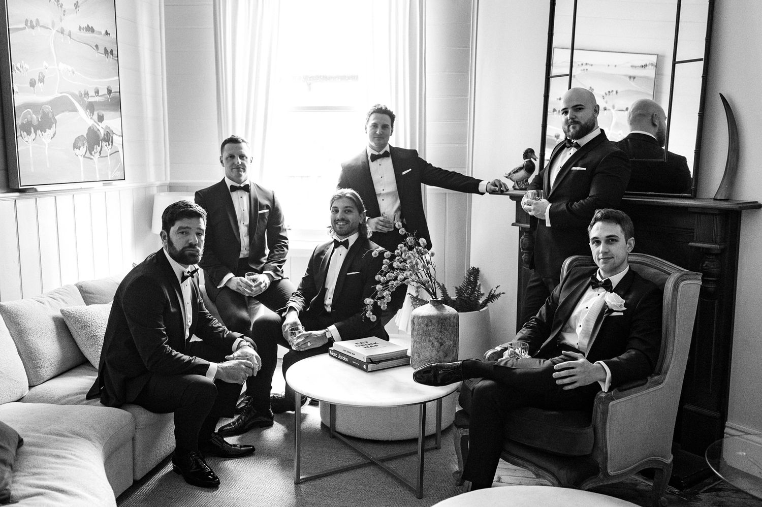 A group of groomsmen in black tuxedos gather on elegant white couches in a bright room for pre-wedding photos.