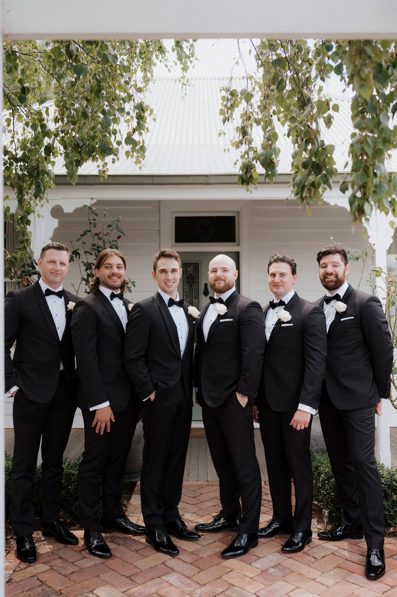 Groomsmen in matching black tuxedos and bowties pose together on a white porch with overhanging greenery.