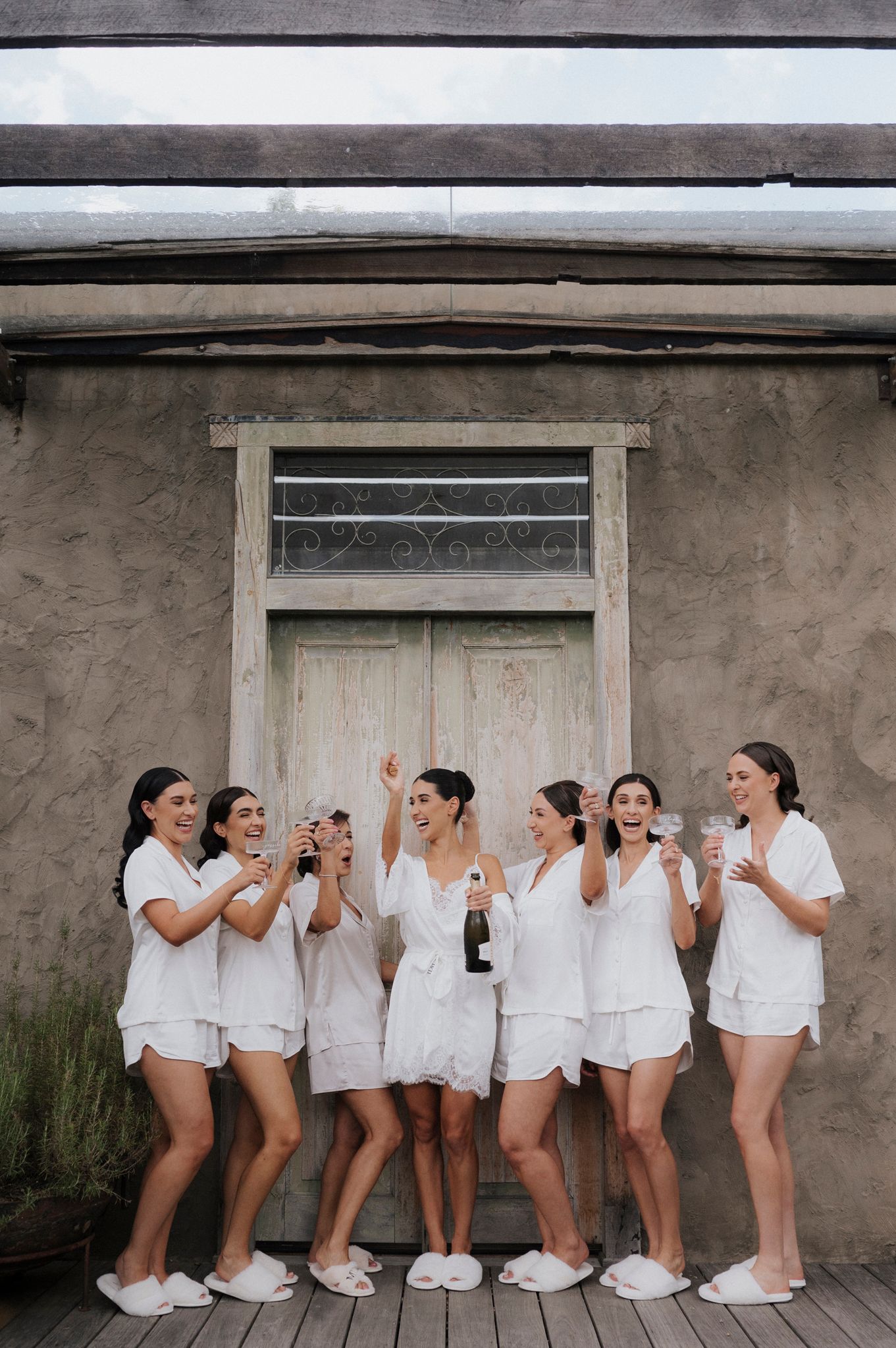 A bridal party in all-white outfits poses together against a rustic wall.