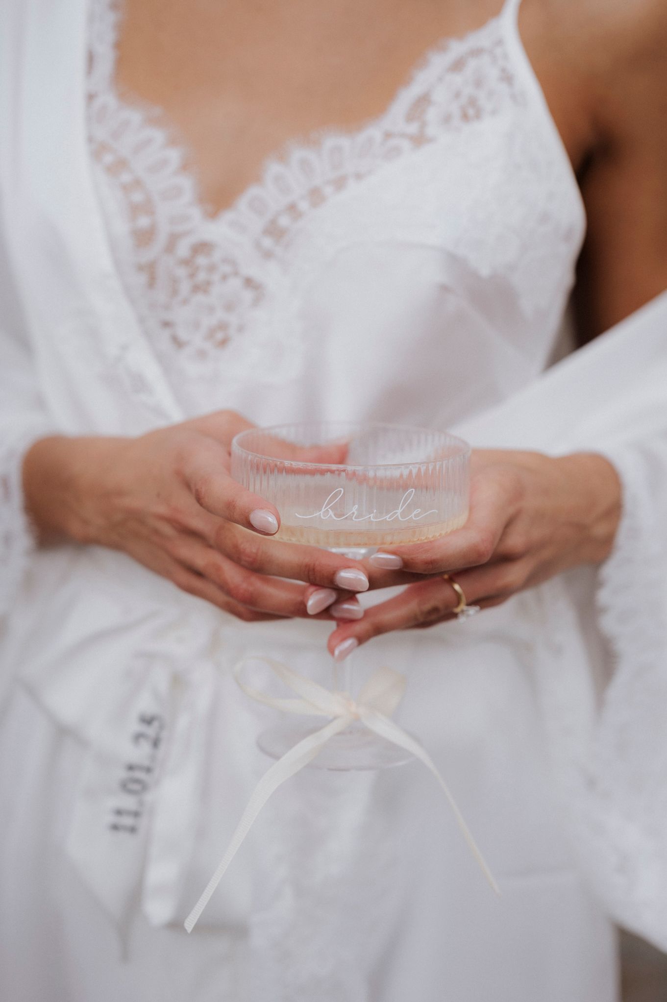 Hands holding a delicate crystal glass with 'Bride' text, wearing a white lace wedding dress.